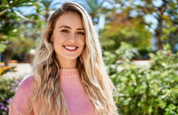 A woman with long blonde hair wearing a pink sweater stands outdoors, smiling at the camera with greenery and sunlight in the background.