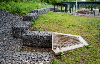 Outdoor drainage system featuring gravel, stone steps, and a metal grate. The system is part of a modern urban park with walking paths and green spaces, showcasing effective water management.
